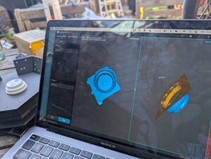 laptop screen with two 3d images of a bottle cap rotated in different directions, behind it, the actual object on a turntable platform