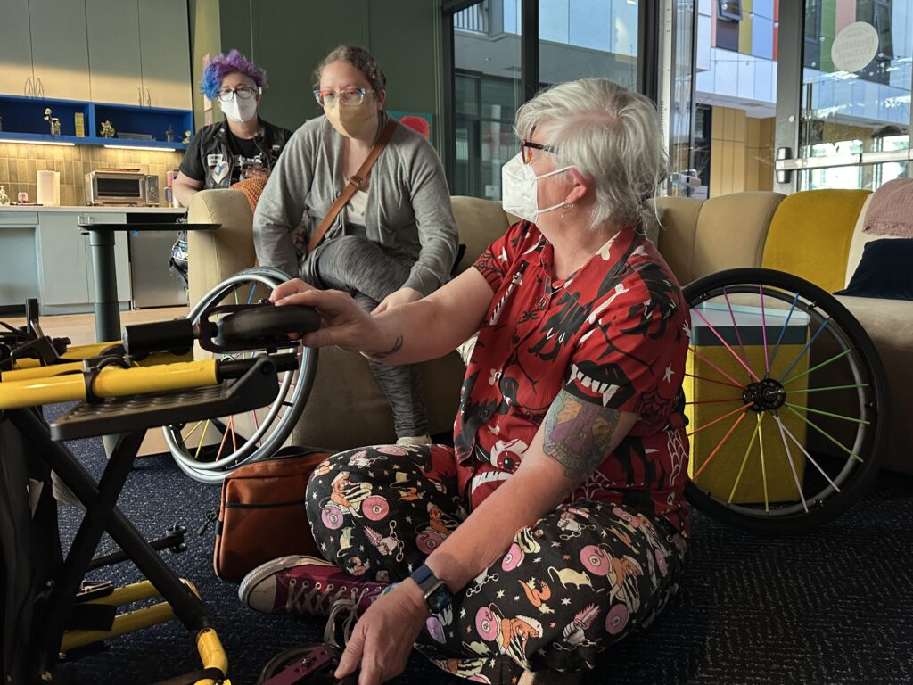woman wearing a mask with tools working on a manual wheelchair with its rider consulting