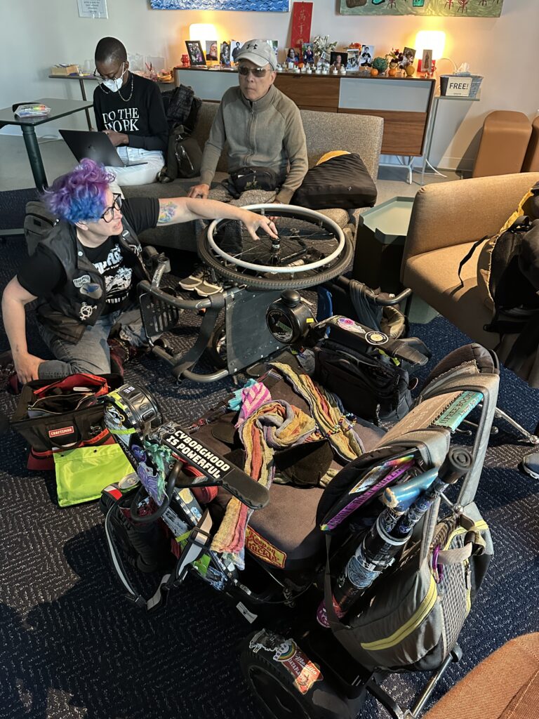 overhead shot of liz on the floor with the manual chair and its stuck wheel, empty powerchair in foreground