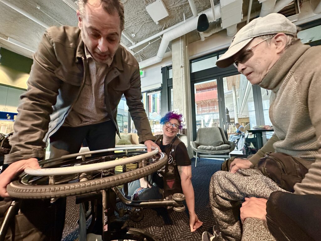 Vince Lopez bending over a stuck wheel with its owner bending over to see the problem, liz laughing a bit in background