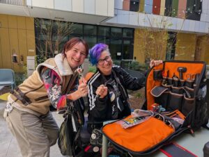 Liz in powerchair, Olga standing, smiling together as they display a large backpack open to show wheelchair fixing tools