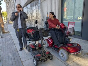 Two guys looking cool on the sidewalk, one on a big mobility scooter and the other standing in sunglasses on the phone