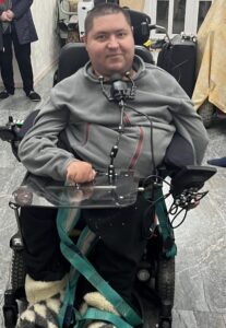a dark haired man in a grey hoodie riding a powerchair, smiling