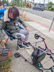 olga on the sidewalk, bending over a partially disassembled rollator with tools at their side