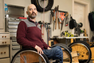 a white man with beard seated in manual chair, in a workshop full of tools