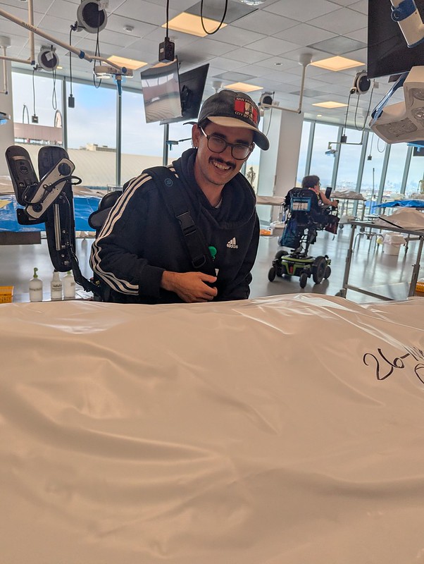 a man in a powerchair smiling over a table with, presumably, a body in a body bag draped in a sheet