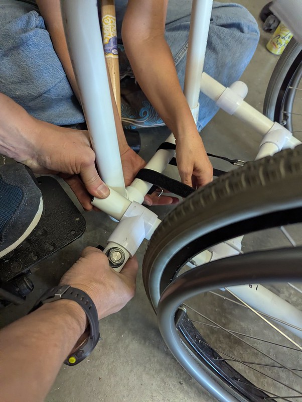 three people's hands pulling together a pvc pipe wheelchair