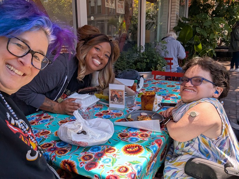 three people smiling around a colorful table of food outside