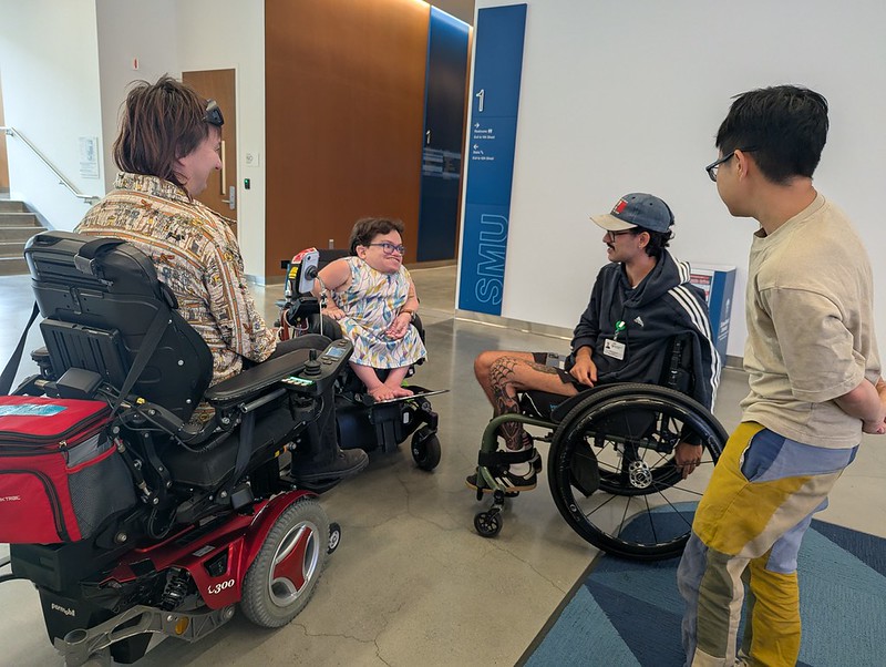 four people in a college building lobby, three in wheelchairs