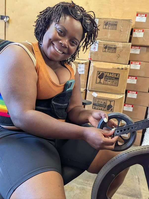 a black non binary femme appearing person smiling with tools in hand over a wheelchair caster