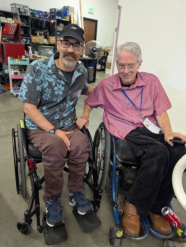 Two manual wheelchair users pose together, arms and wheels linked