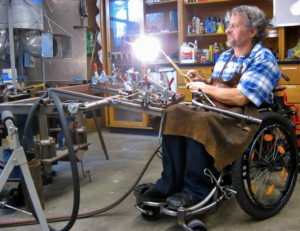 a white man with gray hair seated in a manual wheelchair in a workshop
