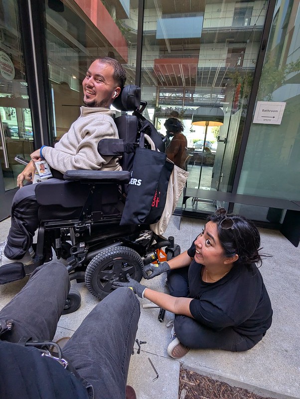 man in powerchair with a woman on the floor next to his rear wheel with tools
