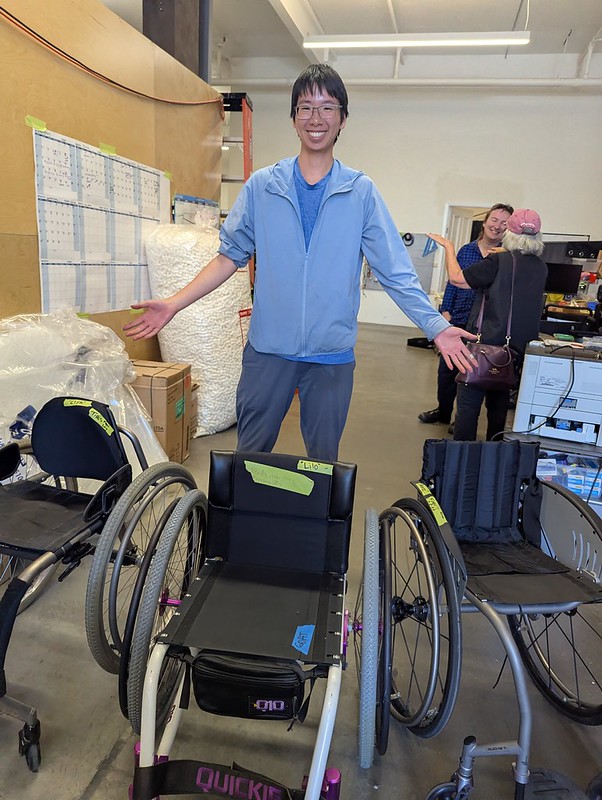 tall brown skinned person spreading arms over three manual wheelchairs