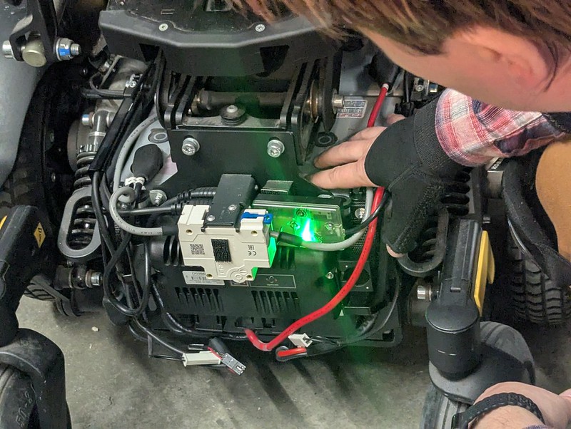 Olga points at battery terminals and compartment on a permobil M3 powerchair